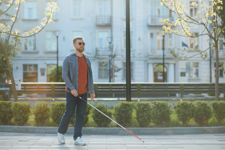 Blind person walking along a sidewalk with a cane.